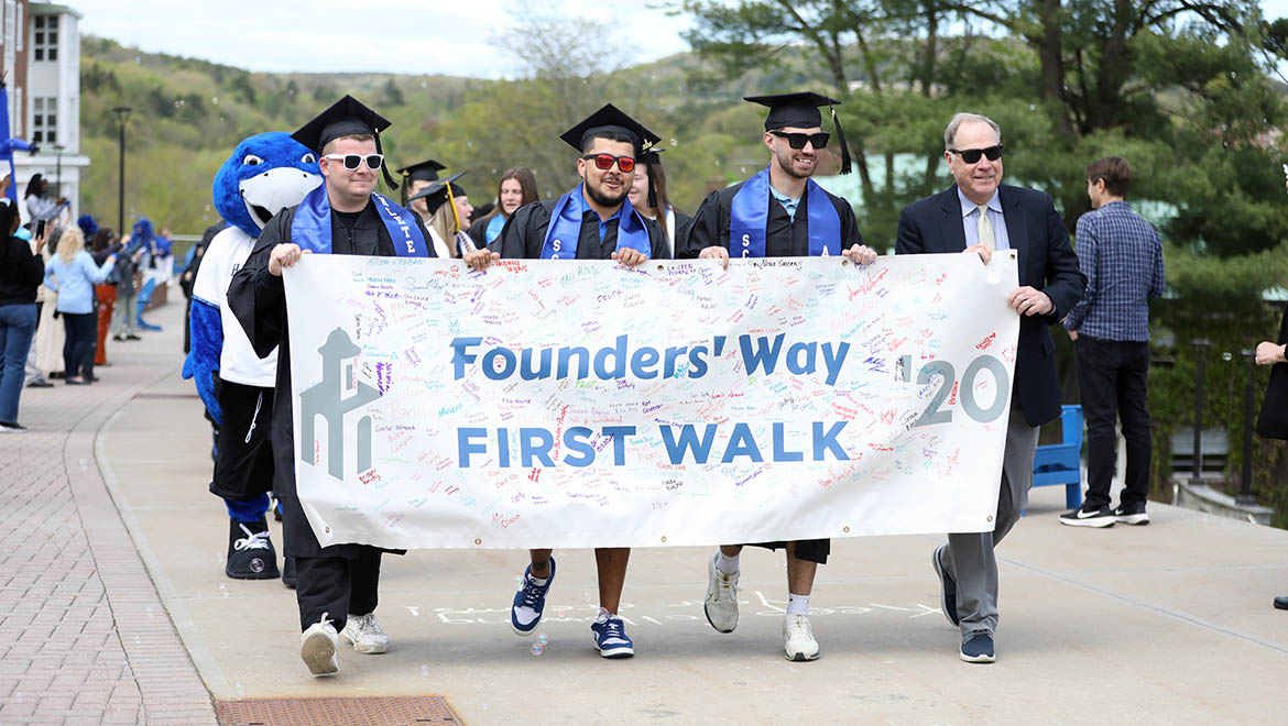 Hartwick College graduates of the Class of 2024 on Last Walk on Founders' Way with Interim President James Mullen holding class banner from First Walk in 2020